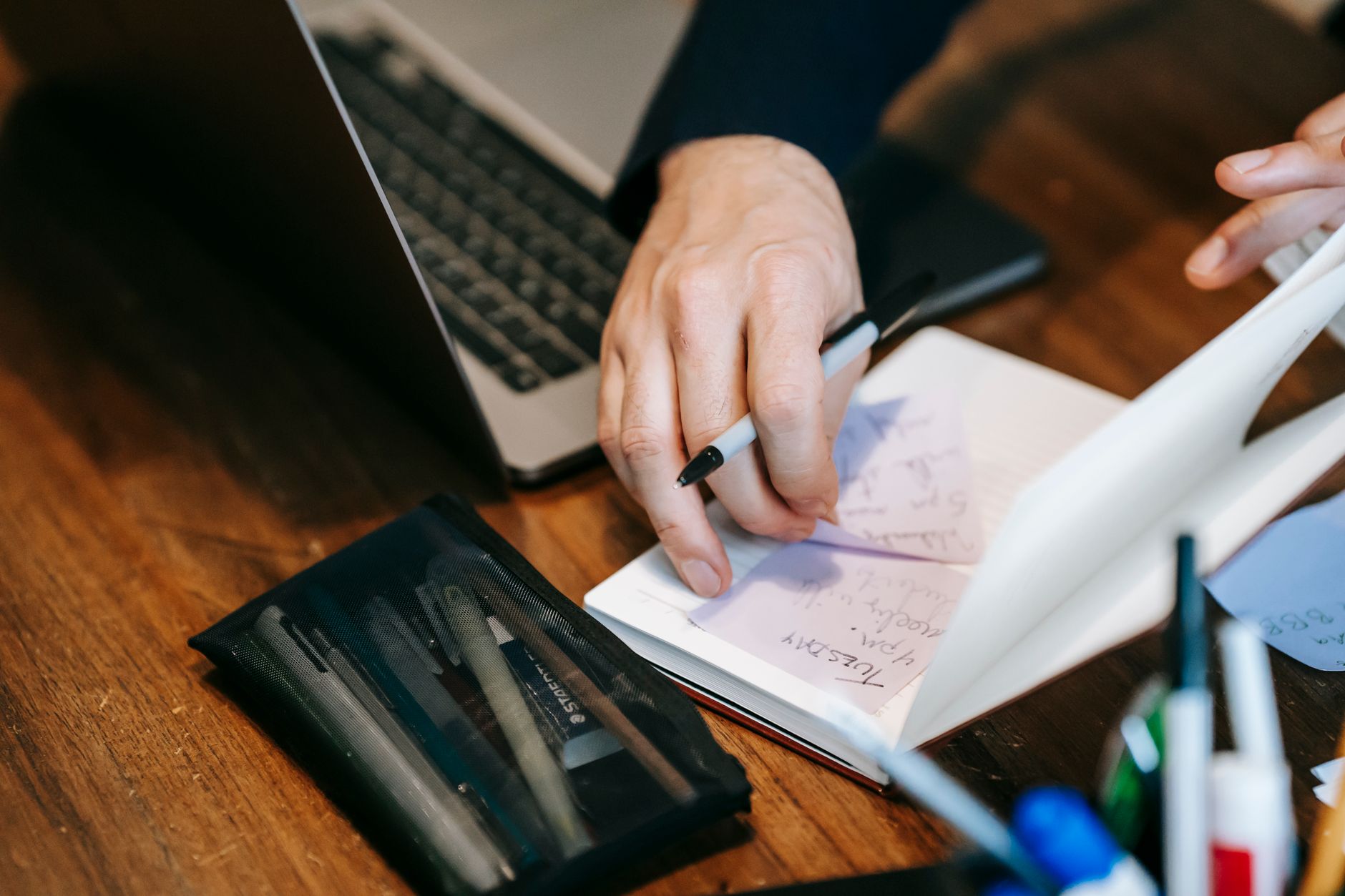 Man's hands writing notes in a notebook beside a laptop on a wooden desk.