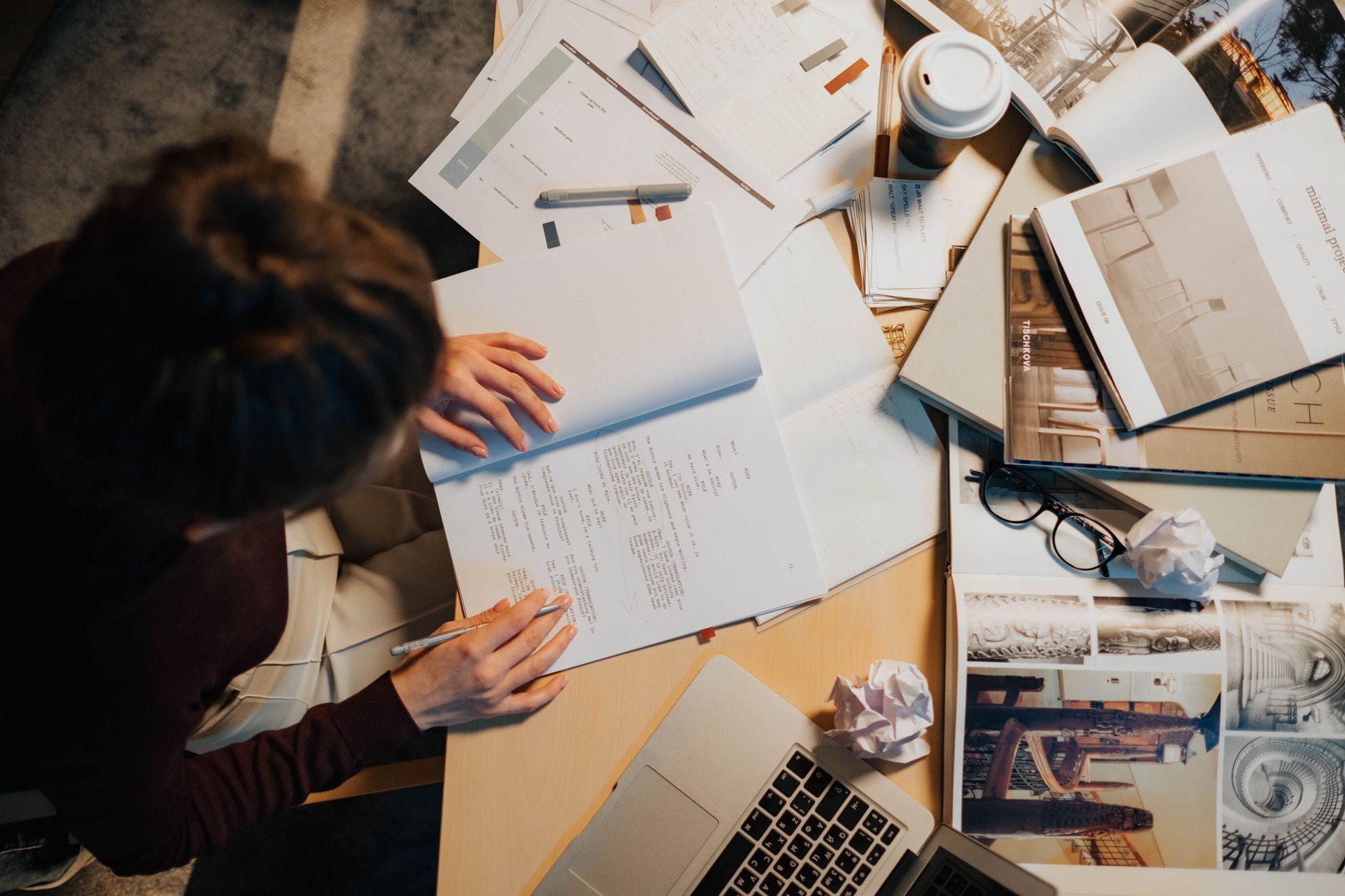 Overhead view of a cluttered work desk with a person writing notes, surrounded by papers, laptop, and coffee cup.