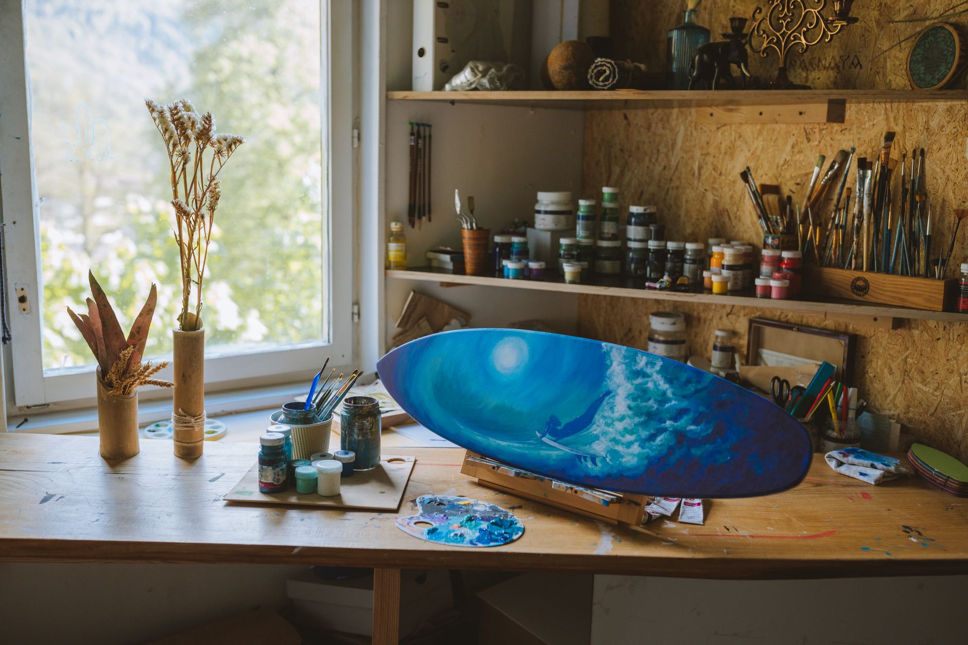 An artistic home studio featuring a vibrant blue ocean painting on a skateboard with art supplies and brushes near a window.