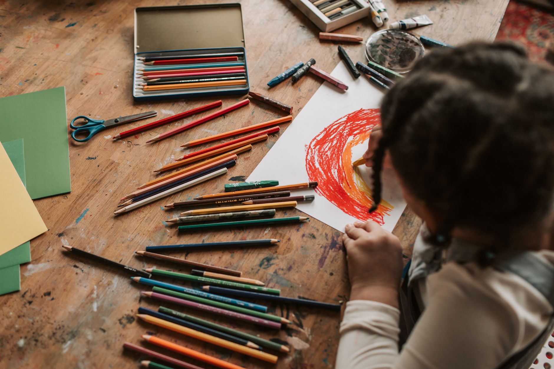 A young child draws a colorful picture with pencils on a wooden table, surrounded by art supplies indoors.