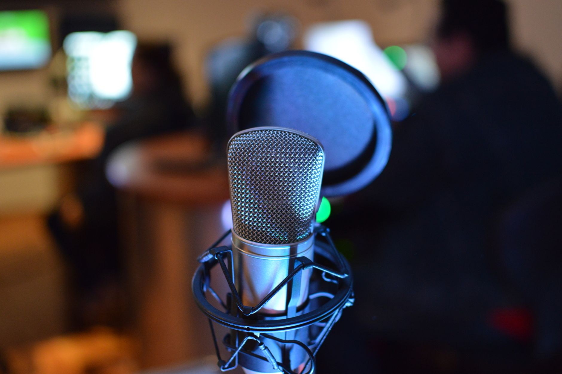 Microphone de studio argenté sur suspension, avec filtre anti-pop, devant un arrière-plan flou de studio éclairé par des écrans.