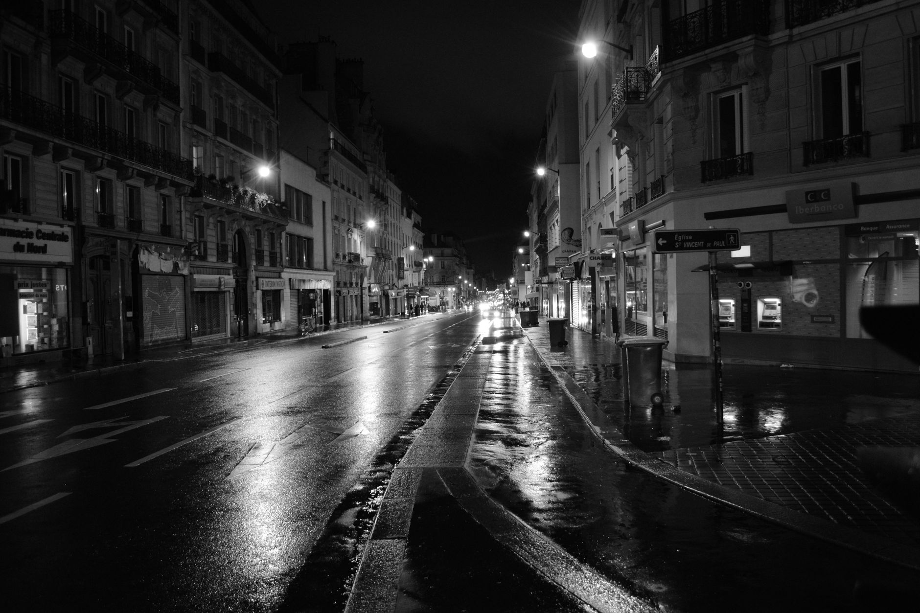 Rue vide de nuit en noir et blanc, chaussée mouillée reflétant les lampadaires, immeubles anciens à balcons et commerces fermés.