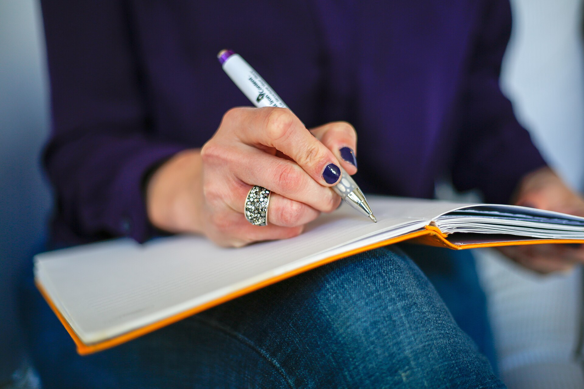 Une main qui écrit au stylo dans un carnet ouvert posé sur les genoux